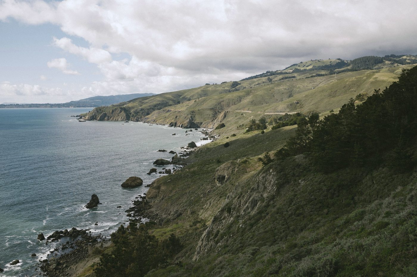 Green coastal hills dropping to a rocky shoreline under broken cloud