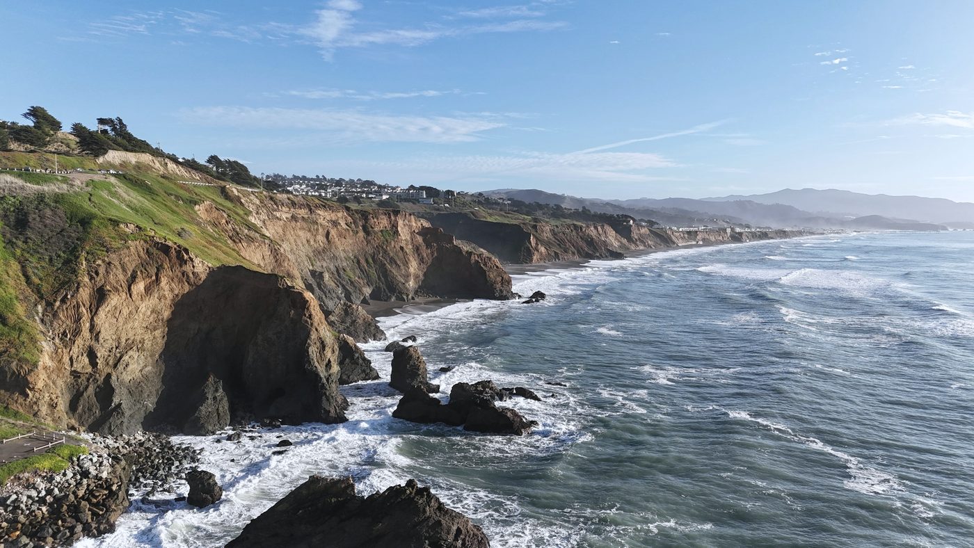 Eroded cliffs above rocks and breaking surf, with houses along the clifftop and hills beyond
